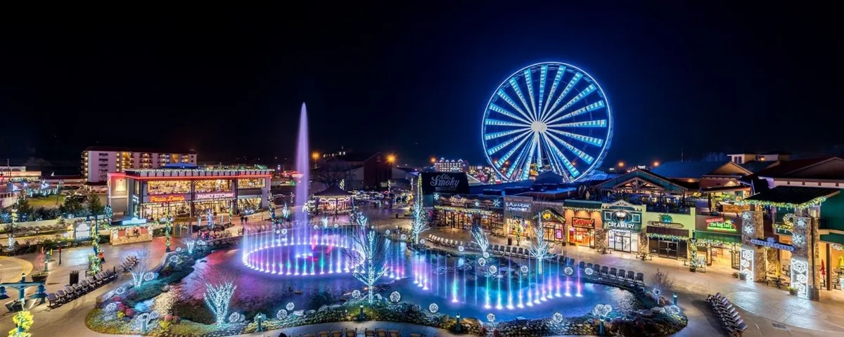 A lively exterior shot of a bustling entertainment plaza, featuring a vibrant fountain, illuminated Ferris wheel, charming shops, and twinkling lights. The scene is alive with opportunities for shopping, dining, and entertainment in the heart of Pigeon Forge.