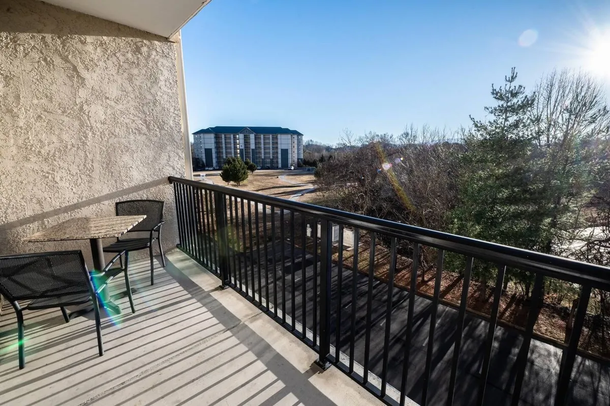A tranquil balcony scene with a sturdy railing, comfortable outdoor furniture, and a distant view of another building surrounded by mature trees. The balcony offers a private, scenic spot ideal for sipping coffee or relaxing as the sun sets.