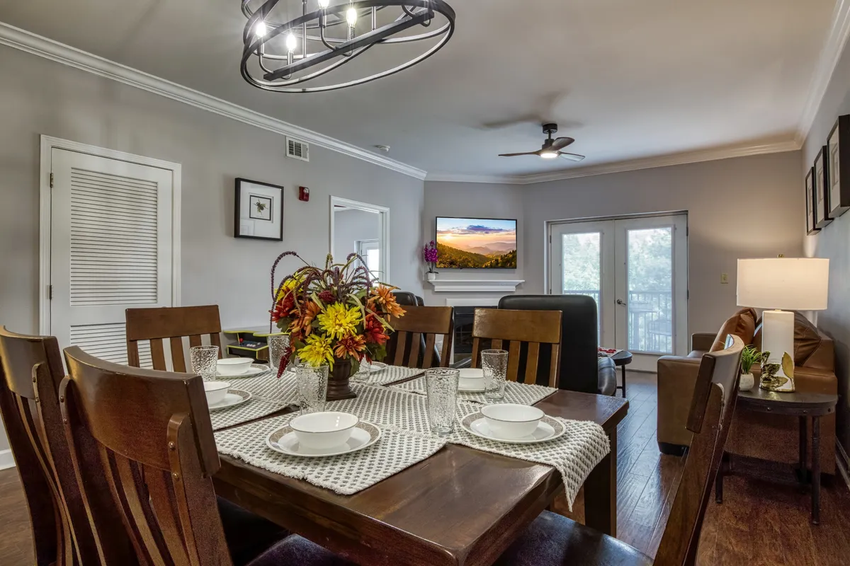 This view captures a dining table set for a meal, a comfortable sofa area, and a television with a scenic backdrop. Decorative touches like a floral centerpiece and set dishes add a sense of occasion and warmth, hinting at celebrations and shared meals.