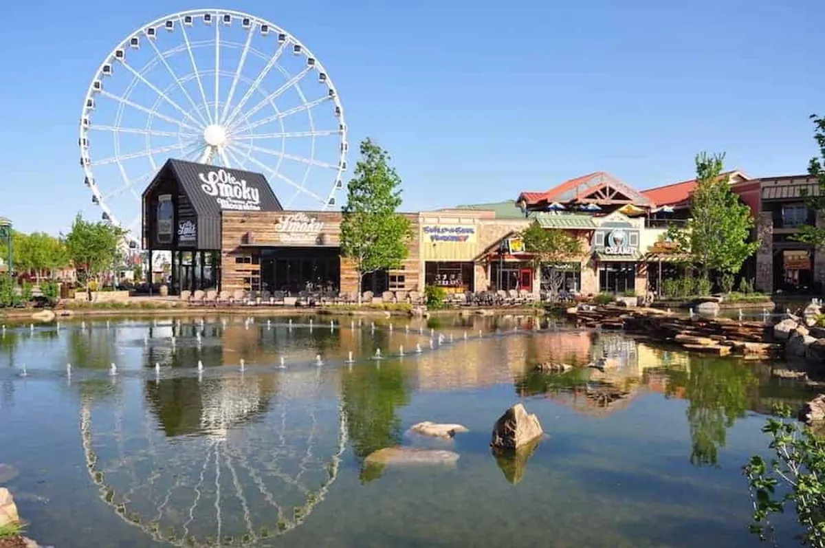 This image captures a lively scene at The Island in Pigeon Forge, featuring the iconic Ferris wheel, sparkling water feature, and bustling storefronts. Families and visitors can be seen strolling along, creating an atmosphere of excitement and community engagement.