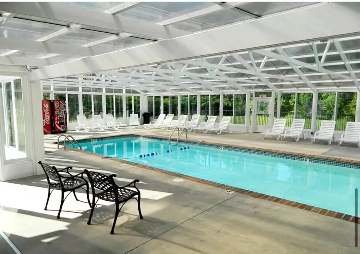 A bright indoor swimming pool area featuring sparkling water, several lounge chairs along the poolside, and a Coca-Cola vending machine. Sunlight streams through the windows, creating a relaxed, inviting atmosphere for leisurely swims or lounging together.